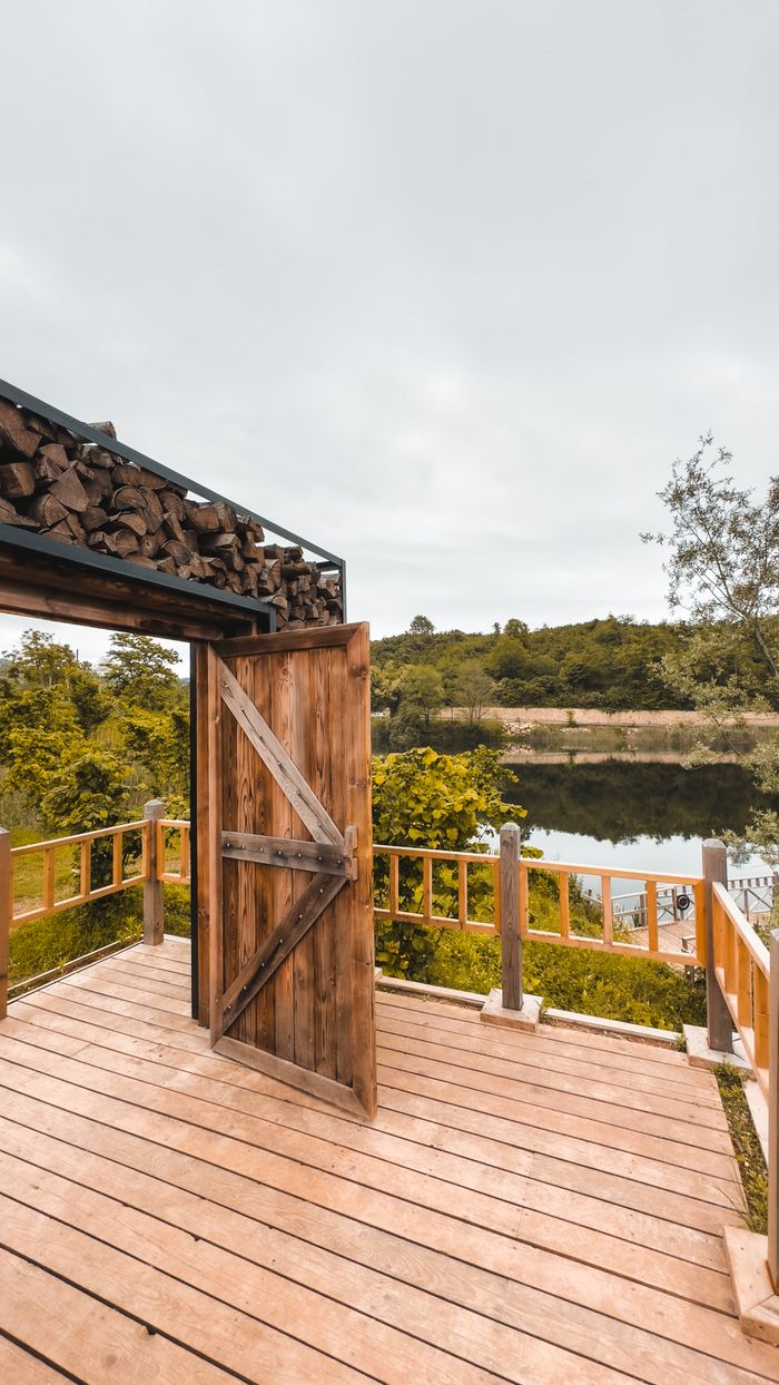A rustic wooden deck with an open door, overlooking a calm lake surrounded by greenery.