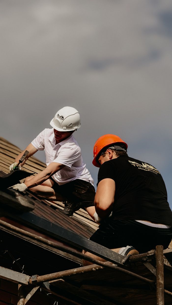 Two construction workers wearing helmets work on a roof under a cloudy sky.